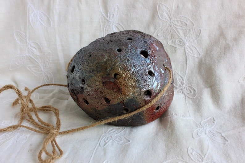 K&ouml;nnte beinhalten: Ein brauner, grauer und goldener Keramik-H&auml;ngepflanztopf mit einem Seilgriff. Der Topf hat eine raue, strukturierte Oberfl&auml;che und ist mit kleinen L&ouml;chern verziert.