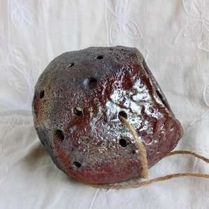 K&ouml;nnte beinhalten: Ein kleiner, brauner Keramiktopf mit einer rauen, strukturierten Oberfl&auml;che und L&ouml;chern f&uuml;r die Drainage. Er hat ein rustikales, handgemachtes Aussehen und h&auml;ngt an einer Schnur.