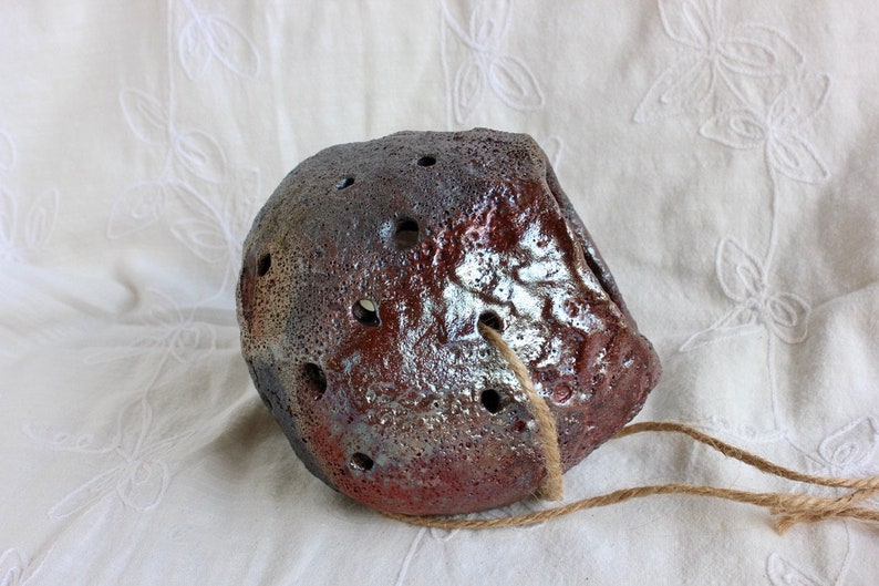 K&ouml;nnte beinhalten: Ein kleiner, brauner Keramiktopf mit einer rauen, strukturierten Oberfl&auml;che und L&ouml;chern f&uuml;r die Drainage. Er hat ein rustikales, handgemachtes Aussehen und h&auml;ngt an einer Schnur.