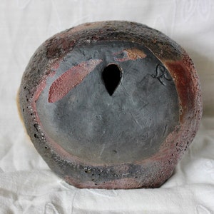 K&ouml;nnte beinhalten: Eine schwarze Keramikvase mit einer rauen, strukturierten Oberfl&auml;che. Die Vase hat ein kleines Loch in der Mitte des Bodens und eine rotbraune Glasur an einigen Stellen.