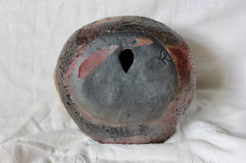 K&ouml;nnte beinhalten: Eine schwarze Keramikvase mit einer rauen, strukturierten Oberfl&auml;che. Die Vase hat ein kleines Loch in der Mitte des Bodens und eine rotbraune Glasur an einigen Stellen.