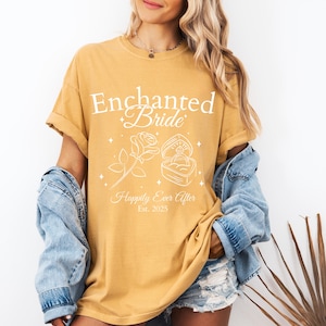 May include: Mustard yellow t-shirt with the text "Enchanted Bride" and "Happily Ever After Est. 2025". The design includes a rose and a ring in a box. The shirt has rolled-up sleeves.
