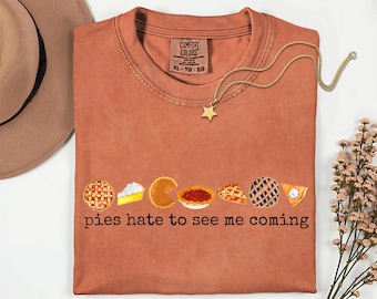 Può includere: Una maglietta Comfort Colors color ruggine con una grafica di torta e la scritta "pies hate to see me coming". Una collana d'oro con un ciondolo a stella è drappeggiata sulla maglietta. La maglietta è taglia XL.