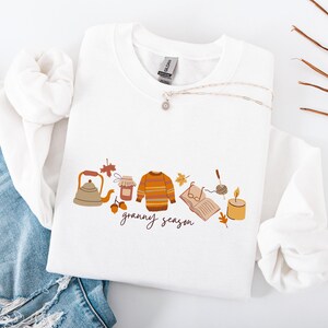 Può includere: Felpa bianca con grafica a tema autunnale. Il design include una teiera, un barattolo di marmellata, un maglione a righe, un libro, una candela e la scritta "granny season". È visibile anche una collana d'argento con un ciondolo.