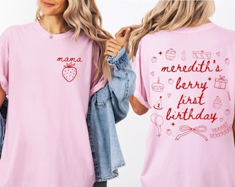 May include: Light pink t-shirts with red graphics. One shirt has "mama" and a strawberry. The other shirt says "meredith's berry first birthday" with birthday-themed illustrations. The shirts are likely for a first birthday celebration.