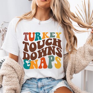 May include: White t-shirt featuring the text "TURKEY TOUCHDOWNS & NAPS" in a retro font. The text is in various colours, including brown, orange, teal, and yellow. The shirt is a casual, comfortable style.