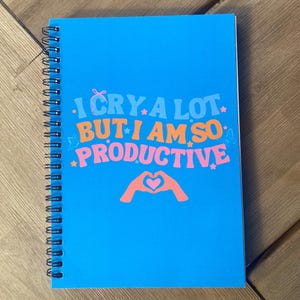 May include: A spiral-bound notebook with a bright blue cover. The cover features the text "I CRY A LOT BUT I AM SO PRODUCTIVE" in orange, pink, and white. A graphic of hands forming a heart is below the text. The notebook is on a wooden surface.