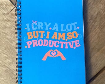 May include: A spiral-bound notebook with a bright blue cover. The cover features the text "I CRY A LOT BUT I AM SO PRODUCTIVE" in orange, pink, and white. A graphic of hands forming a heart is below the text. The notebook is on a wooden surface.