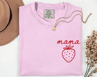 May include: A light pink t-shirt with the word "mama" in red script and a red strawberry graphic. A gold necklace with a star pendant rests on the shirt. The shirt has a Comfort Colors tag.