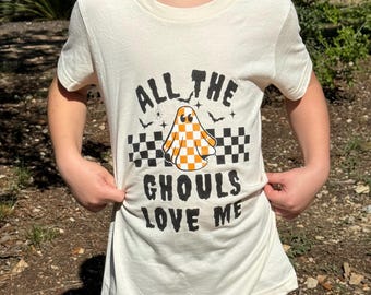 Può includere: T-shirt color crema con la scritta nera "ALL THE GHOULS LOVE ME". La maglietta presenta un fantasma dei cartoni animati con un motivo a scacchi arancione e bianco. Pipistrelli e ragni sono inclusi nel design. La maglietta è indossata da una persona.
