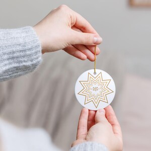 May include: A white, round ornament with a gold starburst design. The ornament is held by a gold string and features a smaller, detailed design in the center. The overall aesthetic is clean and elegant, suitable for holiday decor.