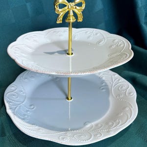 May include: Two-tiered serving stand with scalloped edges. The plates are light gray with a pink trim and embossed floral designs. A gold-colored bow tops the central support. Ideal for displaying desserts or appetizers.