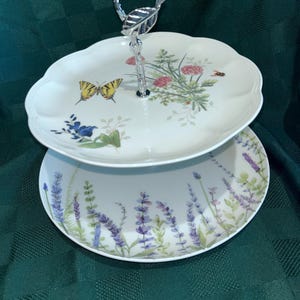 May include: A two-tiered serving tray with a silver handle. The top tier features a butterfly and floral design, while the bottom tier displays lavender sprigs. The tray is ideal for serving desserts or appetizers.