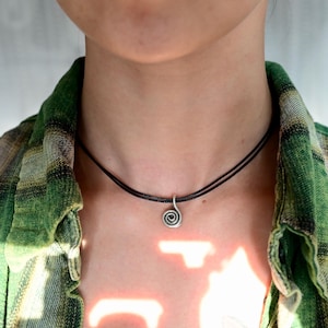 Puede incluir: Un collar con un colgante de espiral plateado en un cordón negro. El collar lo lleva una persona con una camisa de cuadros verde y negro.