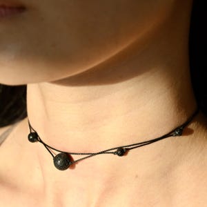 Könnte beinhalten: Eine schwarze Choker-Kette mit drei kleinen schwarzen Lavasteinen. Die Kette besteht aus dünnem schwarzem Garn.