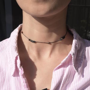 May include: A delicate black beaded necklace with small, dark-coloured beads. The necklace is worn against a pink and white striped button-down shirt. The beads are spaced along a thin black cord, creating a minimalist design.