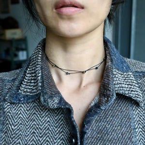 Puede incluir: Primer plano de una persona que lleva un collar gargantilla negro con pequeñas cuentas doradas y negras. El collar está en capas y tiene un diseño delicado y minimalista. La persona lleva una camisa con estampado azul y blanco.