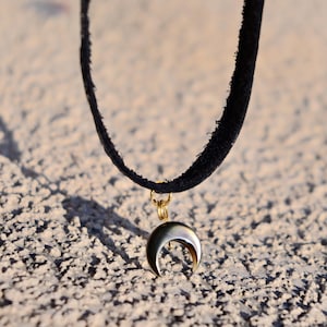 Puede incluir: Un collar gargantilla de ante negro con un colgante de media luna dorada.