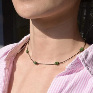 Puede incluir: Un collar sencillo con un cordón negro y cinco cuentas de jade verde. El collar lo lleva una persona con una camisa de rayas rosa y blancas.