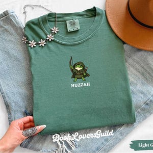 Puede incluir: Una camiseta verde salvia con un bordado de una rana vestida de arquero con arco y flecha, acompañada del texto HUZZAH. La camiseta se muestra junto a unos vaqueros azul claro, un sombrero marrón y un collar de flores.