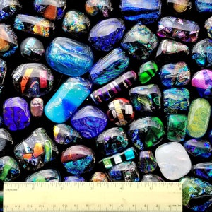 May include: A collection of dichroic glass pieces in various shapes and sizes, displaying a spectrum of colors including blues, greens, purples, and golds. A ruler is at the bottom, indicating the size of the glass pieces. The glass has a reflective, iridescent quality.