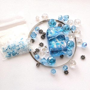 May include: A collection of craft supplies including a rectangular blue glass pendant with a swirling design, clear and blue faceted beads, small silver-colored metal accents, and a bag of small seed beads. The items are arranged on a white surface.