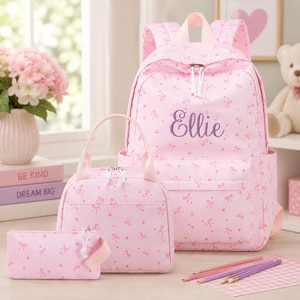 May include: A light pink backpack, lunch bag, and pencil case set featuring a repeating pattern of small bows and hearts. The backpack is personalized with the name Ellie embroidered in purple script on the front pocket, with white zippers.