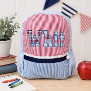 May include: A seersucker backpack featuring a red and white vertical stripe pattern on the upper section and a light blue and white stripe pattern on the lower section. The name Whit is embroidered in a blue plaid fabric across the center.