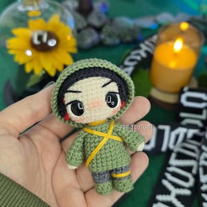 Pode incluir: Uma pequena boneca feita à mão em crochê, representando uma personagem com cabelo preto, olhos pretos grandes e bochechas rosadas. A boneca usa uma capa verde com capuz, uma túnica verde com tiras cruzadas amarelas e calças verdes com detalhes amarelos. O texto ramygurumi é visível.