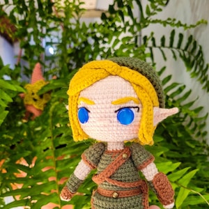 May include: Crocheted figure of a video game character with blonde hair, green clothing, and blue eyes. The figure is holding a sword and is standing on a green surface.