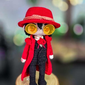 Pode incluir: Boneca de crochê feita à mão vestida com chapéu vermelho, casaco e óculos amarelos. A boneca está vestida com um fato preto e está sobre uma base de madeira. As características da boneca são meticulosamente trabalhadas, mostrando um design único e artístico.