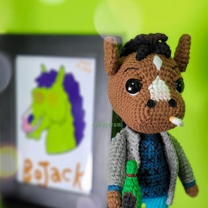 May include: A brown and white crocheted horse plush toy wearing a gray jacket, blue jeans, and a blue and white sweater. The toy is holding a green bottle and has a cigarette in its mouth. The toy is standing in front of a framed picture of a green horse with the word "BoJack" written below it.