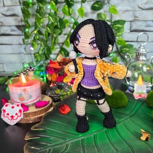 May include: A handmade crochet doll of a singer, wearing a purple top, black skirt, and a yellow jacket. The doll is holding a microphone and has black boots. The doll is standing on a green leaf-shaped base.