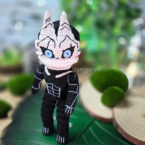 May include: A handmade crochet doll in a black and white color scheme. The doll has a detailed face with blue eyes and a unique design. The doll is standing on a green leaf, with a blurred background of greenery.