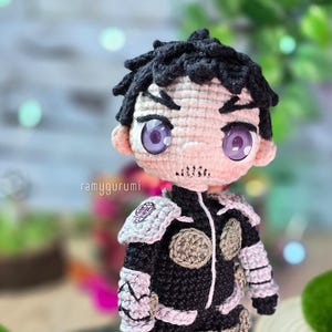 Pode incluir: Uma figura amigurumi de crochê de um personagem com cabelo preto, olhos roxos e um traje preto e cinza. A figura está usando uma armadura com detalhes prateados.