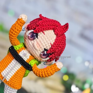 May include: A handmade crochet doll with red hair, large purple eyes, and an orange and white outfit. The doll has a black belt and is posed with its arms raised. The doll's face is detailed with stitched features and small earrings.