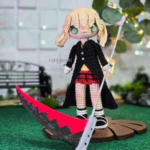 Pode incluir: Boneca de crochê feita à mão de um personagem com cabelo loiro, olhos verdes e um casaco preto. A boneca está segurando uma grande foice vermelha e preta. A boneca está em uma base de madeira.