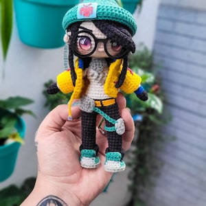 May include: A crocheted amigurumi doll with black hair, wearing a green beanie with a red patch, yellow jacket, black trousers, and green and white trainers. The doll is holding a small, grey bag.