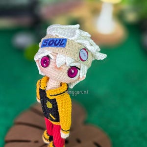 Op de afbeelding: Gehaakte pop met een personage met wit haar, een "SOUL"-patch en grote roze ogen. De pop draagt een gele jas met een zwart vest, een rode broek en geelzwarte schoenen. Hij staat op een houten blad-vormige basis.