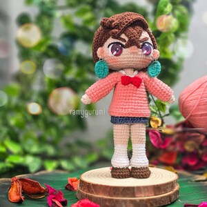 May include: Crocheted amigurumi doll with brown hair, pink sweater, blue skirt, and white socks. The doll has large eyes and is wearing turquoise earrings. The doll is standing on a wooden platform.