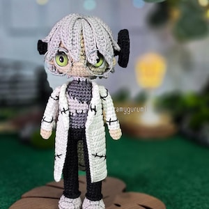 May include: A handmade crochet doll of a character wearing a white lab coat with black stitches, black pants, and a gray sweater. The doll has gray hair, round glasses, and green eyes. The doll is standing on a wooden leaf-shaped base.
