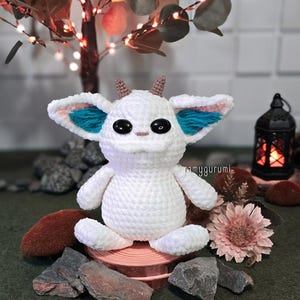 May include: A white crocheted creature with large black eyes, small horns, and teal and pink accents on its ears. The handmade amigurumi is sitting on a small wooden platform, surrounded by rocks and a dried flower. The text "ramygurumi" is visible.