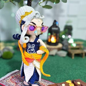 Könnte beinhalten: Handgefertigte Häkelpuppe mit weiß-goldenem Kopfschmuck, Sonnenbrille und einem marineblauen Outfit mit Hasenmotiv. Die Puppe hat einen orangefarbenen Schärpe und steht auf einem Miniaturteppich.