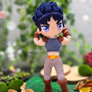 May include: Crocheted amigurumi figure of a character with blue hair, a blue and gray top, brown pants, and brown boots. The figure has a determined expression and is standing with its arms bent at the elbows.