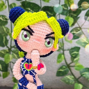 May include: Crocheted doll with green eyes, yellow hair, and a blue and white checkered outfit. The doll is wearing a gold bracelet and a pink butterfly on its arm. The doll is holding its chin with its right hand.