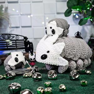Pode incluir: Brinquedos de pelúcia cinza e brancos de crochê com olhos pretos e uma pequena figura são exibidos em uma superfície verde. Sinos de prata de vários tamanhos estão espalhados ao redor dos brinquedos. Os brinquedos têm uma aparência macia e texturizada.