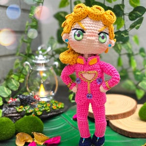 May include: A crocheted doll with blonde hair, green eyes, and a pink outfit. The doll is standing on a green leaf.