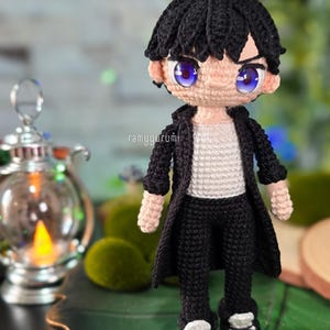 Może przedstawiać: Ręcznie robiona szydełkowa lalka przedstawiająca męską postać z czarnymi włosami, ubraną w czarny płaszcz, spodnie i buty. Lalka ma duże niebieskie oczy i białą koszulę. Lalka stoi na tle rozmytego zielonego tła.