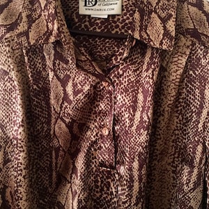 May include: A brown and tan silk blouse with a snake print pattern. The blouse has a button-up front and a collar. The label on the blouse reads "Da Rue of California" and "www.darue.com".
