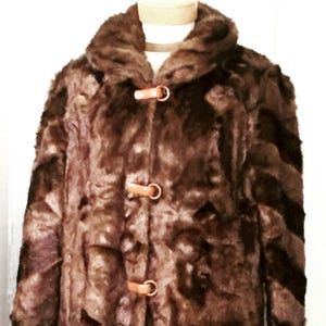 May include: A brown faux fur coat with a wide collar and three leather toggle closures. The coat is displayed on a mannequin and has a textured appearance. The coat is a medium length.